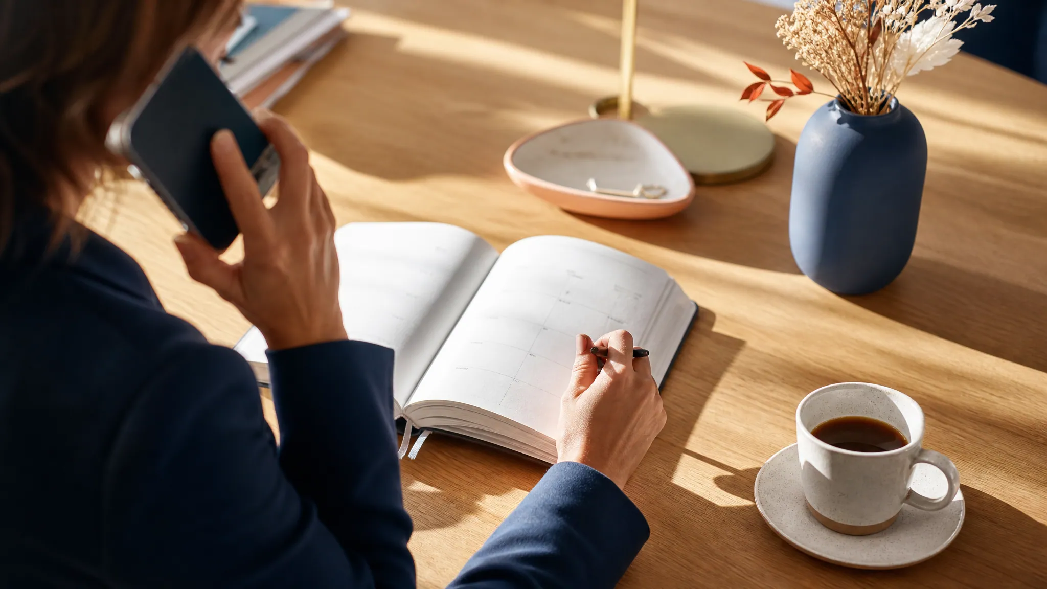 Une main tient un téléphone en pleine conversation près d’un agenda ouvert, sur un bureau ensoleillé