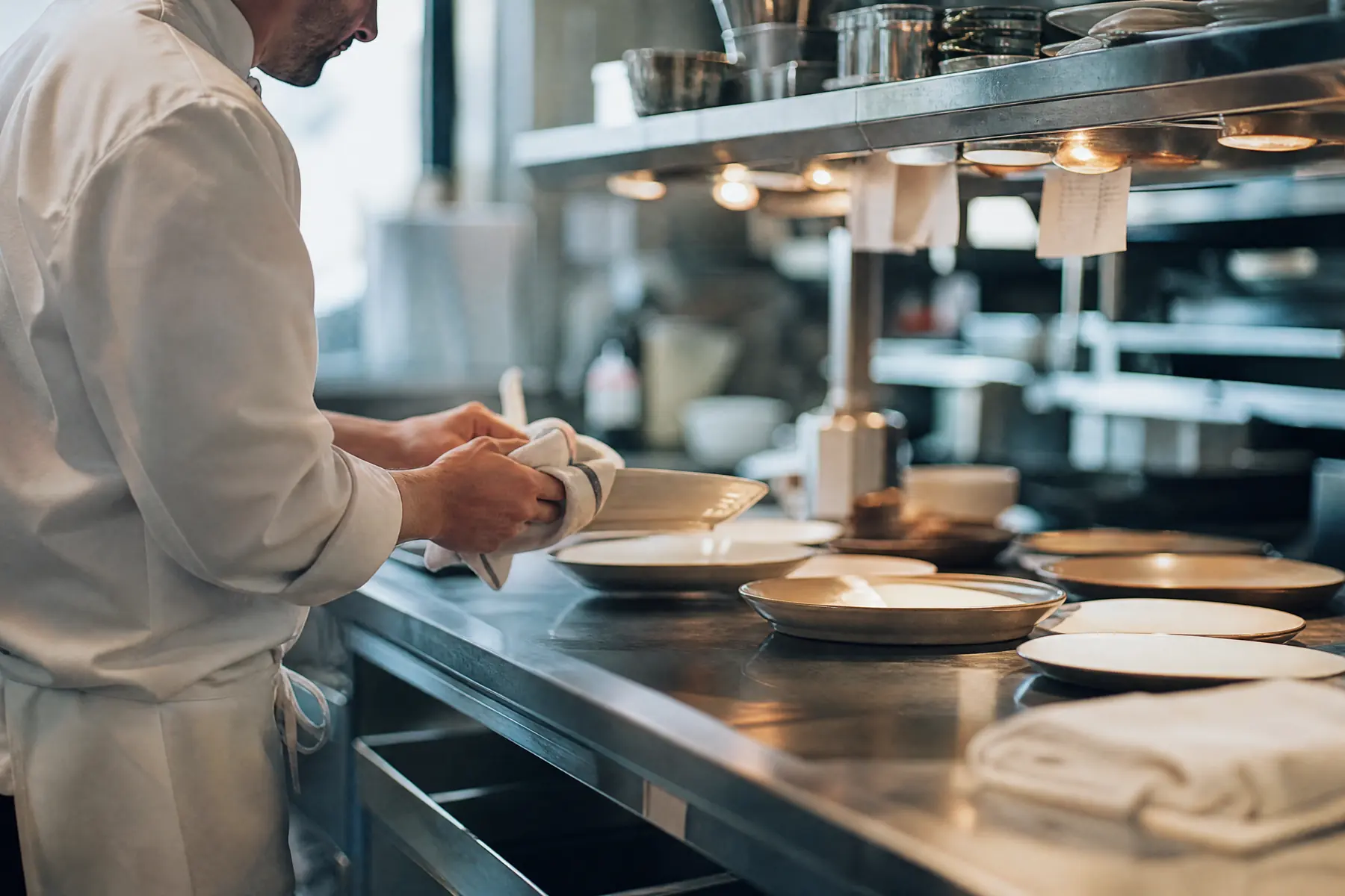Passe d’une cuisine de restaurant en inox pendant la mise en place, un cuisinier en veste blanche essuyant le bord d’une assiette