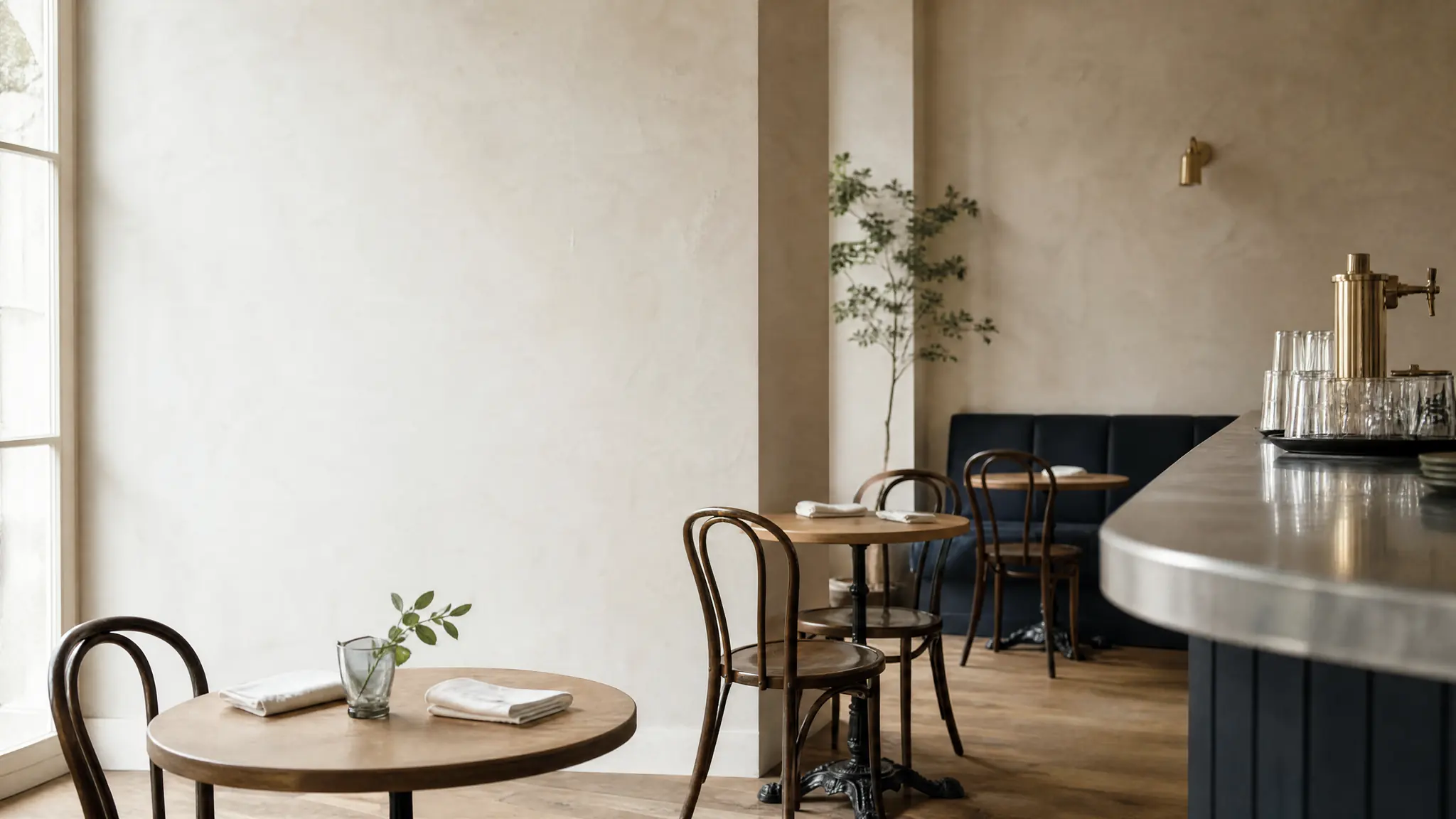 Salle d’un restaurant de quartier avant le service : chaises en bois cintré aux tables rondes, comptoir en zinc en retrait, lumière du jour entrant par une grande fenêtre